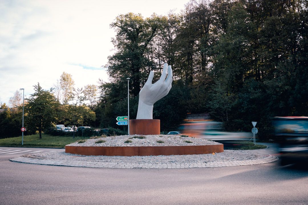 Kreiselskulptur Aarberg: offene Hand aus Mörtel auf Stahlsockel, Skulptur fotografiert mit Langzeitbelichtung und verwischtem Verkehr